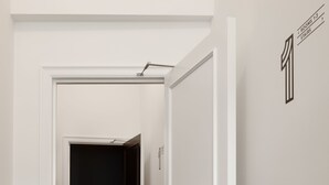 A hallway with a carpet featuring geometric patterns, a white door with a black handle, and a wall sign indicating "ROOMS 1-3 STAIRCASE". - Riga Suites Antwerp (Antwerp)