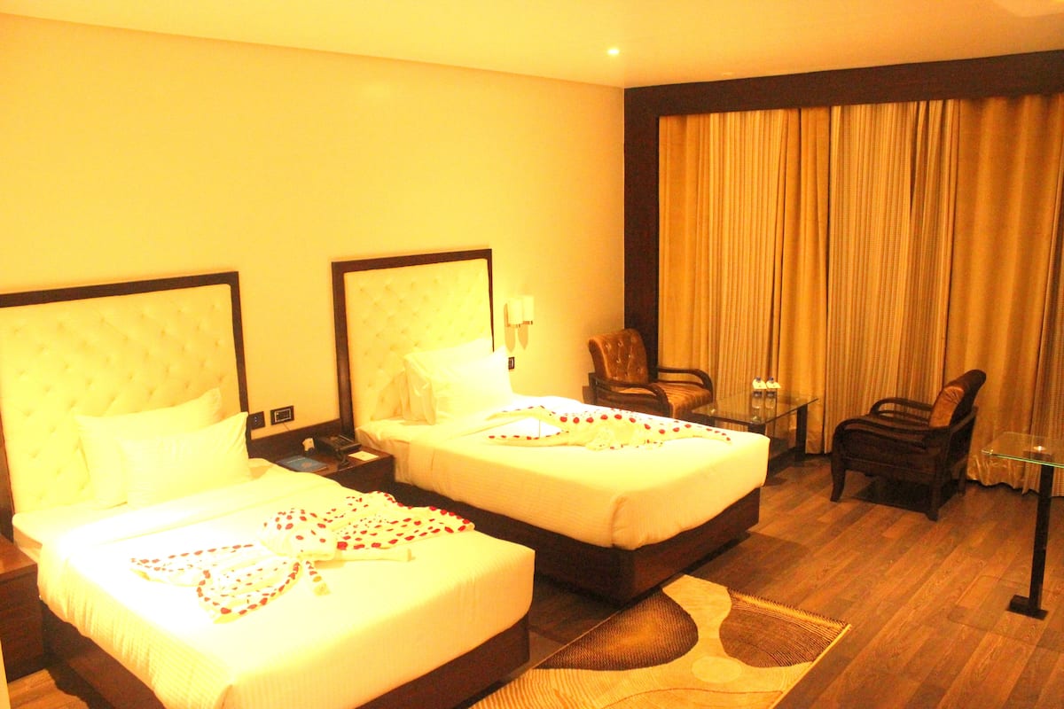 Habitación de hotel con dos camas, una zona de estar con una silla y una mesa, y un ventanal con cortinas.