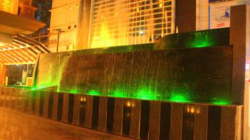 Un elemento acuático con una cascada iluminada en verde, un banco de piedra y un edificio con un letrero para una cocina urbana al fondo.