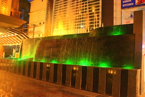 A water feature with a green-lit waterfall, a stone bench, and a building with a sign for an urban kitchen in the background. - Vennington Court (Raipur)