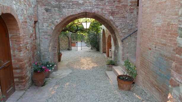 Un pasillo de ladrillo con arco, piso de grava, plantas en macetas y una verja que conduce a un jardín.