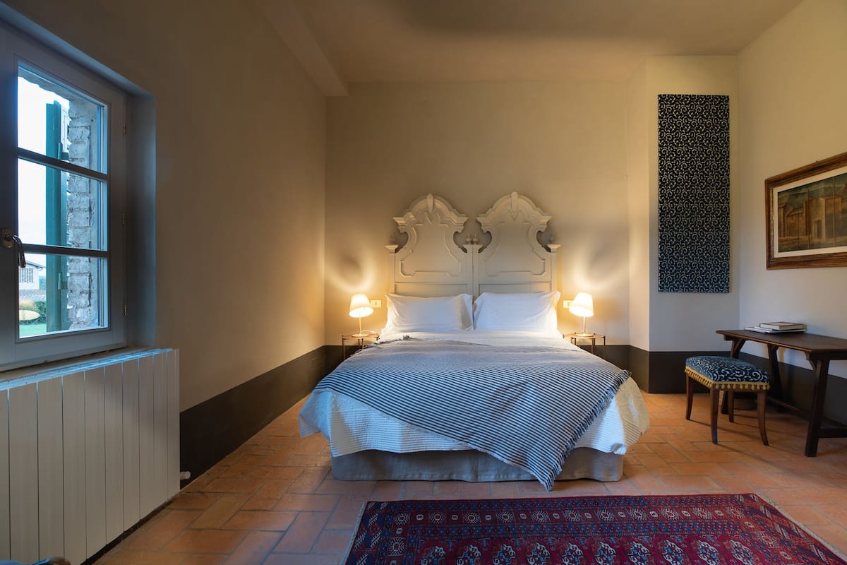 A hotel room with a large bed, two bedside lamps, a small table, a chair, a patterned rug, and a painting on the wall.
