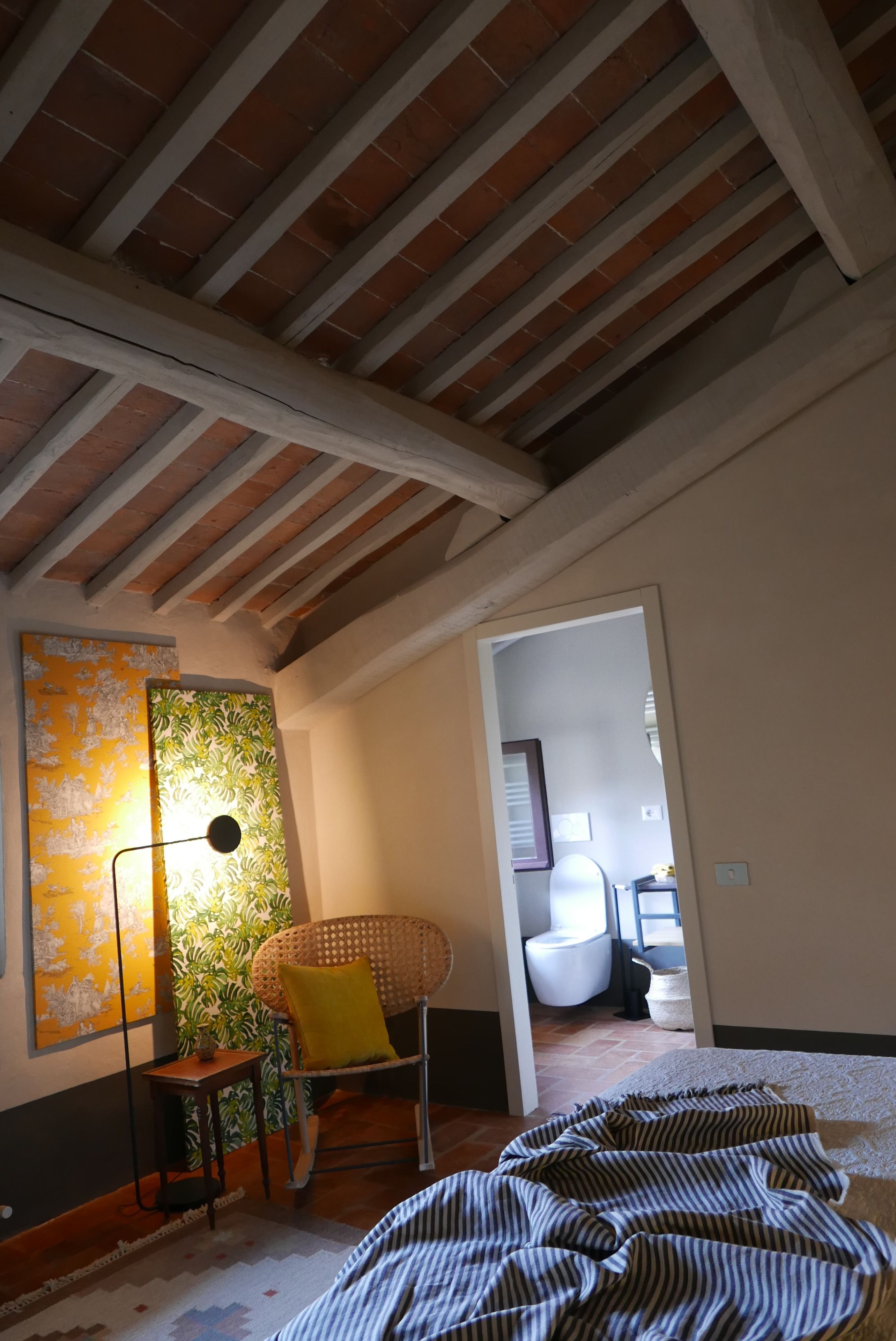 A room with exposed wooden beams, a large abstract painting, a wicker chair with a yellow cushion, and a striped blanket on the floor.