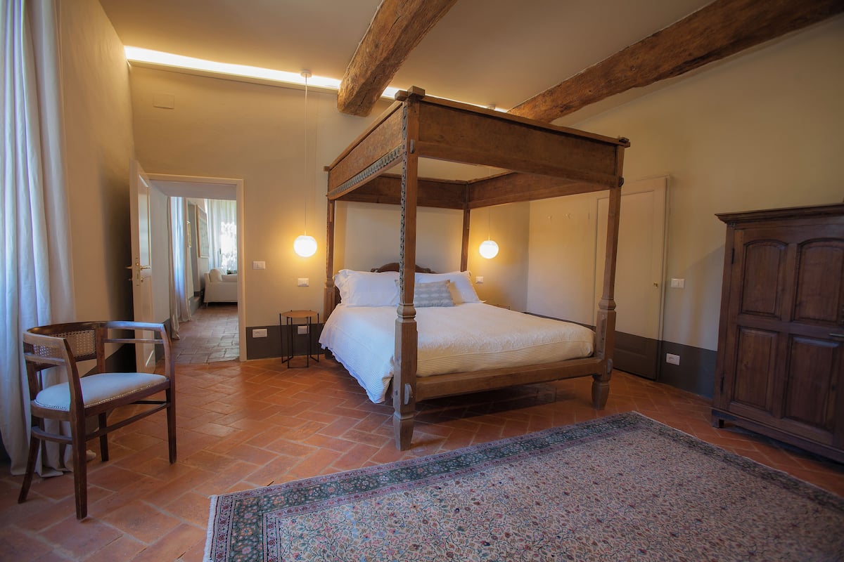 A four-poster bed with a wooden frame, a chair, a cabinet, and a patterned rug in a room with tiled flooring and exposed wooden beams.