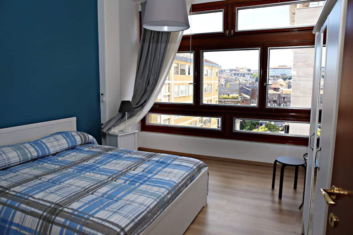 Un dormitorio con una cama, una ventana con cortinas, una silla y vistas a edificios.