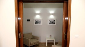 Una habitación con puerta de madera, un sofá color beige, una mesita blanca con un florero y dos lámparas fijadas a la pared.