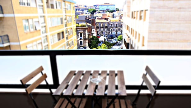 Un balcón con mesa y sillas de madera, con vistas a un paisaje urbano que incluye edificios y coches estacionados.
