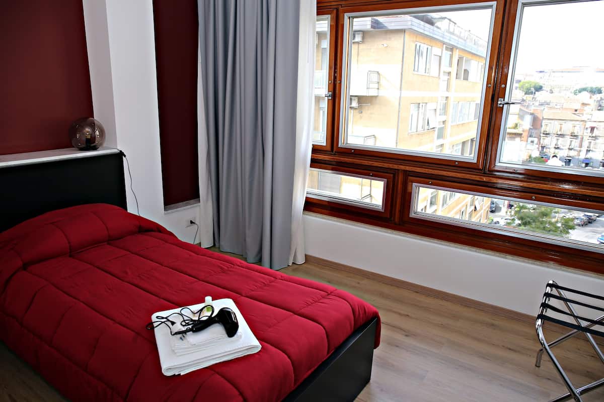 Un dormitorio con una cama roja, una ventana que da a edificios y una mesita de noche con una lámpara.
