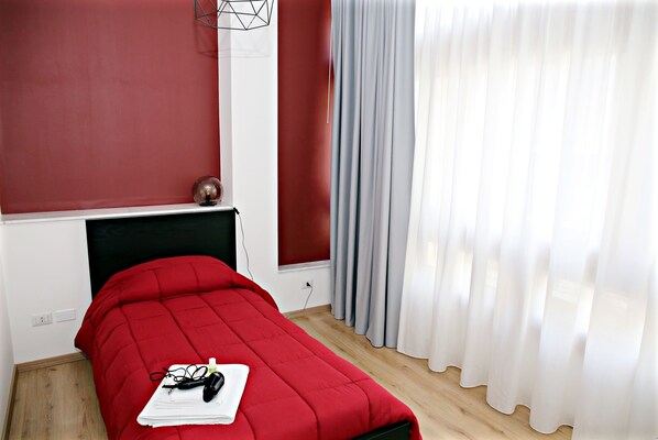 A neatly made bed with a red bedspread, a small table with a lamp, and a window with curtains. - Tosca B&B (Catania)