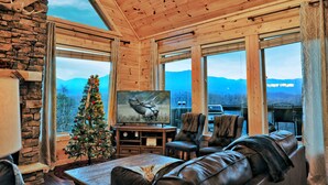 A cozy cabin interior with a large window, a Christmas tree, leather furniture, and a television displaying a moose. - STUNNING Luxury Cabin! Game Room! 4 Fireplaces! Huge Decks Hot Tub! AMAZING View (Gatlinburg)