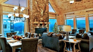 A spacious living area with a stone fireplace, leather furniture, and large windows offering a mountain view. - STUNNING Luxury Cabin! Game Room! 4 Fireplaces! Huge Decks Hot Tub! AMAZING View (Gatlinburg)
