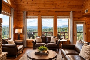 A cozy cabin interior with a fireplace, leather sofas, a round coffee table, and a balcony with a scenic mountain view.