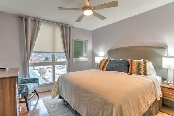 Un dormitorio con cama, escritorio, silla, mesita de noche, lámpara, ventilador de techo y ventana con cortinas.