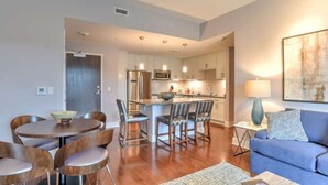 A modern kitchen with stainless steel appliances, a dining area with wooden chairs, and a blue sofa in a living room.