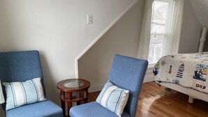 Een kamer met twee blauwe fauteuils met gestreepte kussens, een klein houten bijzettafeltje, een bed met een quilt met nautische motieven, en een trap met een witte leuning.