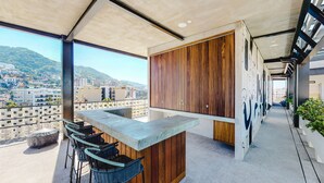 A balcony with a concrete bar and wicker chairs, a wooden cabinet, and a view of a cityscape with buildings and hills. - Beautiful Condo in the heart of Zona Romantica! 2BD Condo for rent in Old Town, (Puerto Vallarta)