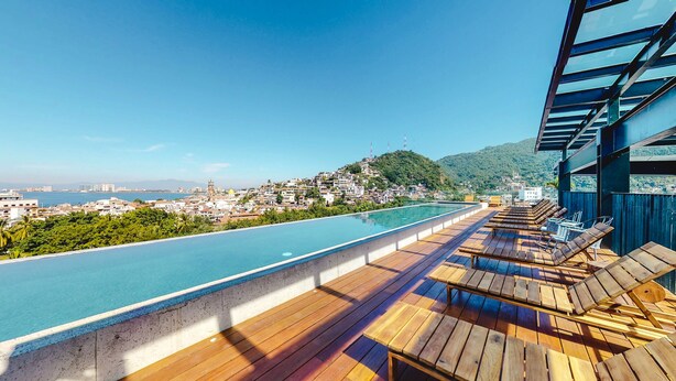 A rooftop infinity pool with wooden deck and lounge chairs, overlooking a coastal cityscape with hills and a clear sky. - Beautiful Condo in the heart of Zona Romantica! 2BD Condo for rent in Old Town, (Puerto Vallarta)