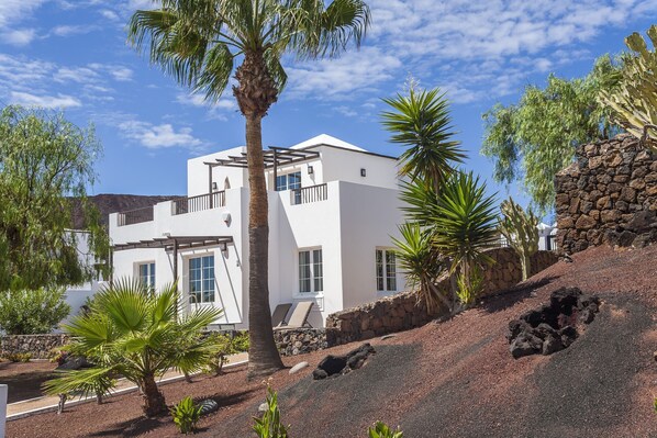 Un edificio moderno blanco con balcón, rodeado de plantas tropicales y una ladera rocosa.