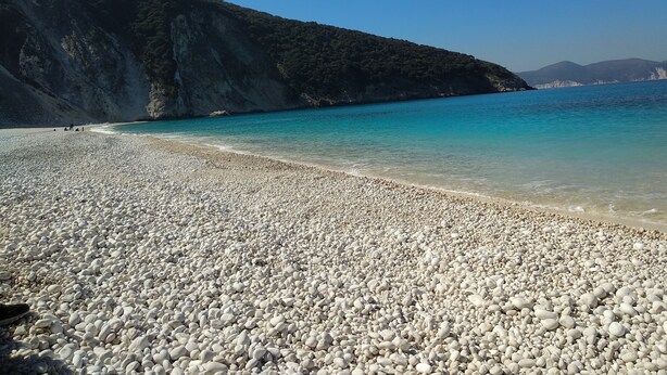 Uma praia de pedras com águas turquesa cristalinas e um cenário montanhoso.
