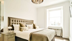 A bedroom with a large bed, a nightstand, a window with a city view, and a chandelier.