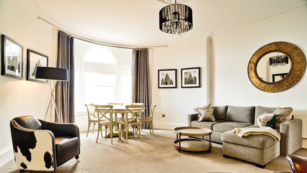 A living room with a sofa, armchair, dining table, and chairs. There is a chandelier and a round mirror on the wall.