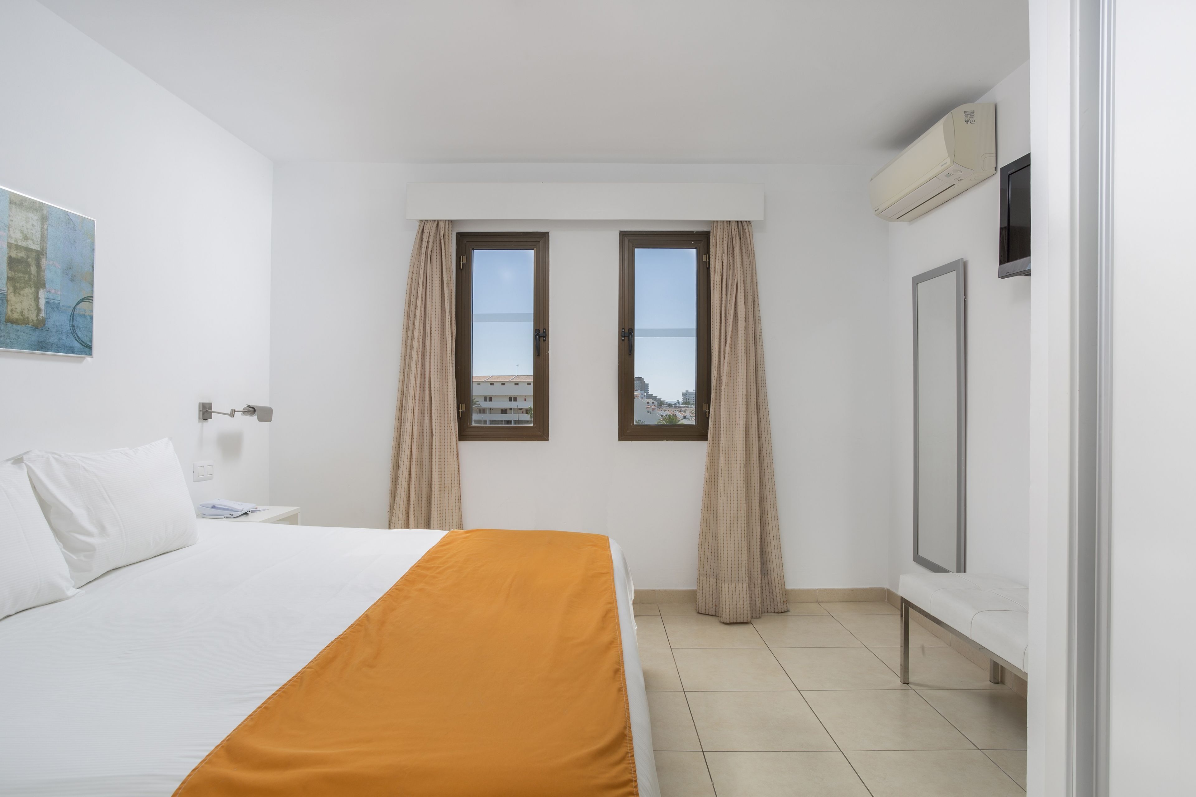 A hotel room with a bed, two windows with curtains, a flat-screen TV, and an air conditioning unit.