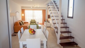 A modern living room with a white sofa, a dining table with fruit, a staircase, and a large window. - 1 Bedroom Apartment, Sunset Bay (Costa Adeje)