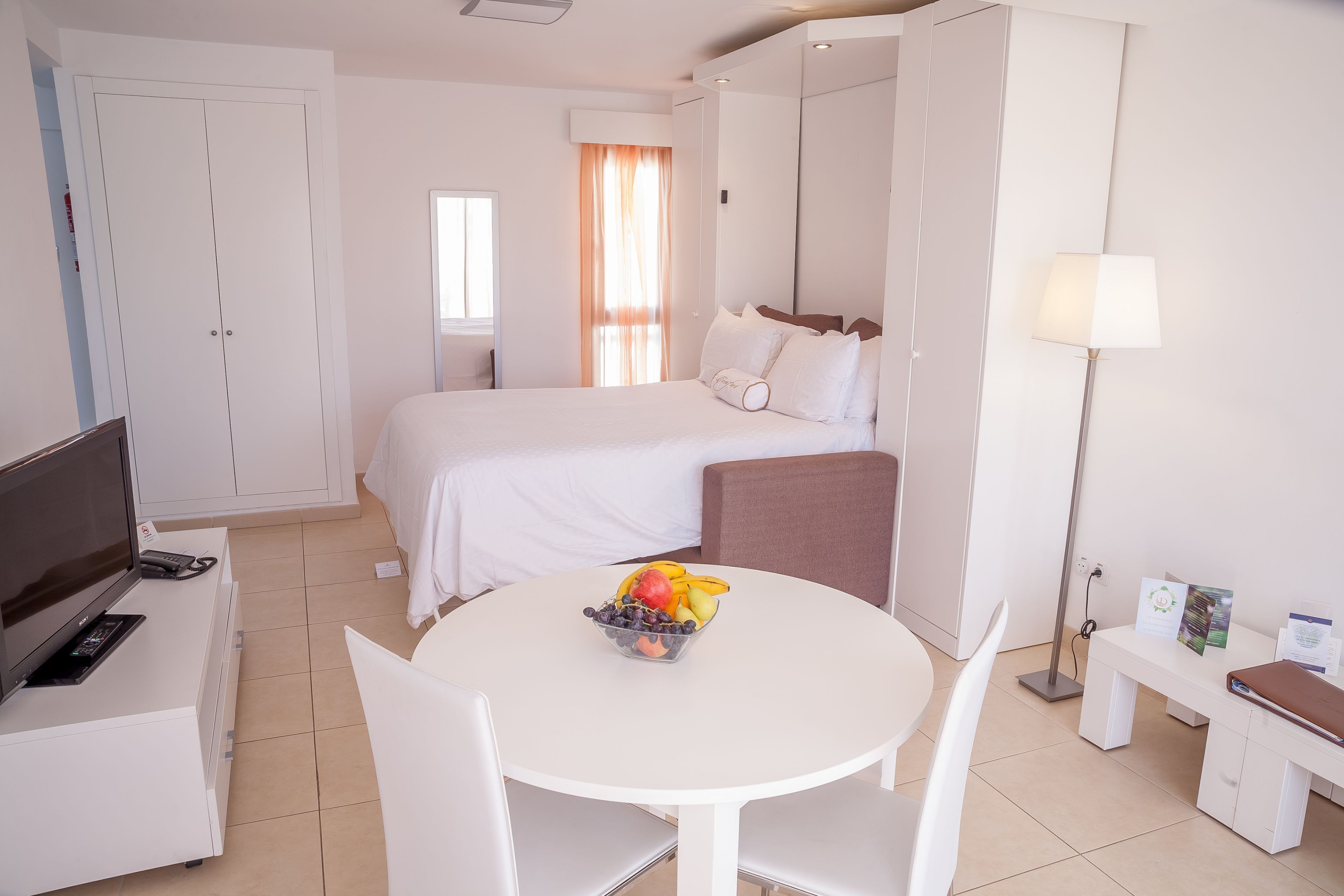 Una camera d'albergo con un letto, una televisione, una scrivania con un computer, un tavolo con della frutta e una lampada.