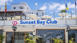 The exterior of Sunset Bay Club with a clear view of the entrance, a signboard, and surrounding buildings. - 1 Bedroom Apartment, Sunset Bay (Costa Adeje)