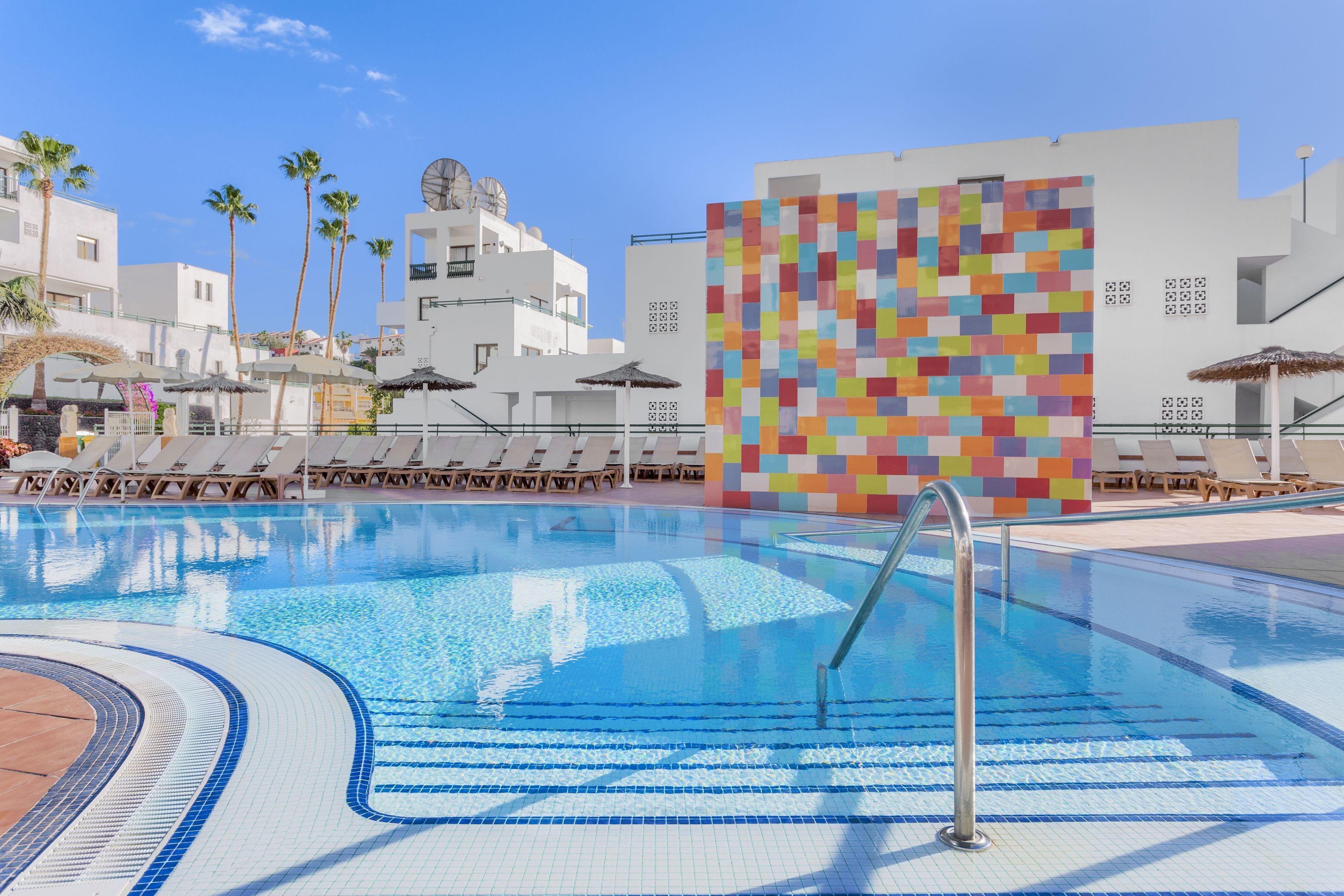 A swimming pool with lounge chairs and umbrellas, surrounded by white buildings and palm trees.