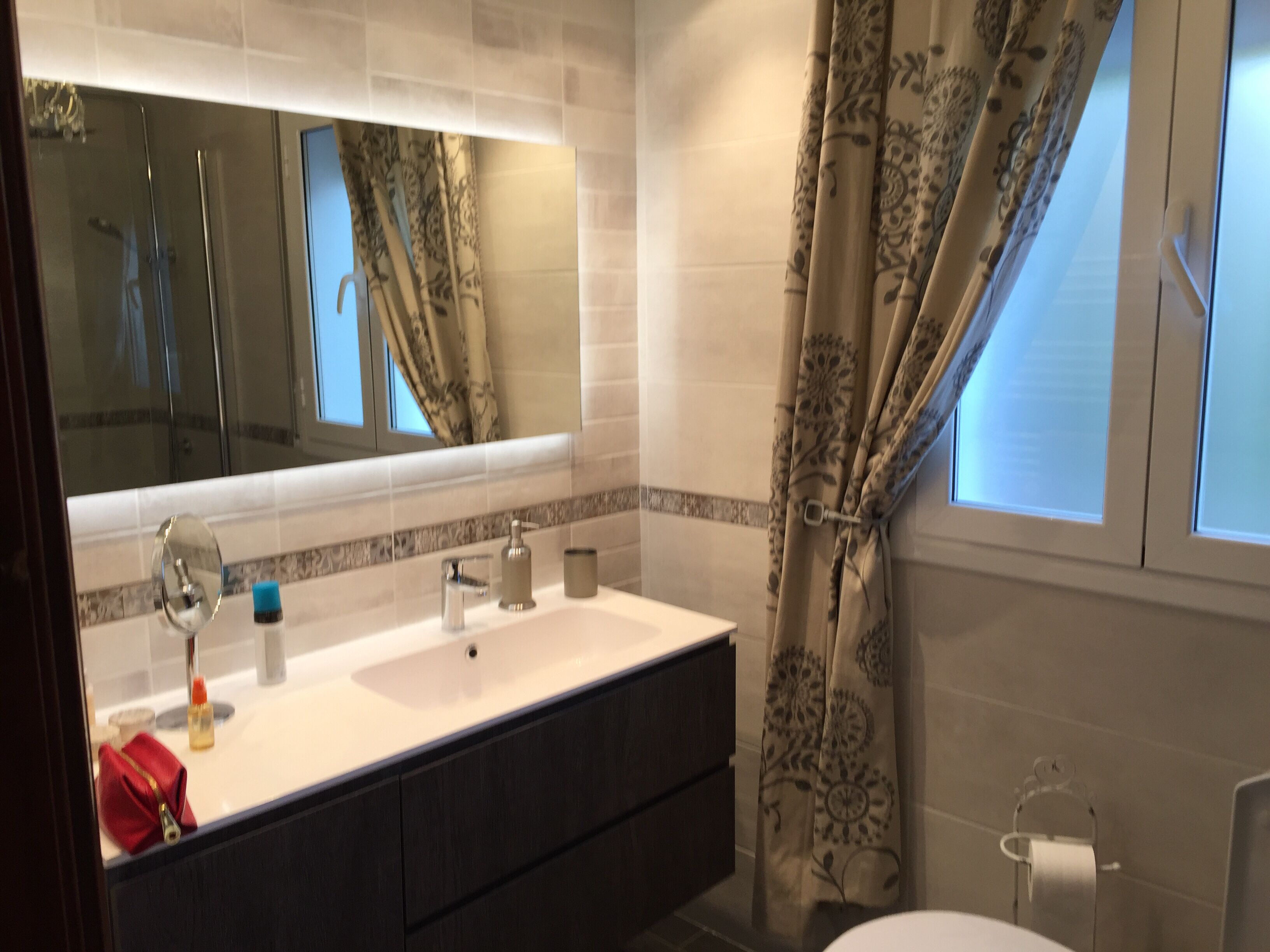 A modern bathroom with a large mirror, two sinks, a toilet, and a shower.