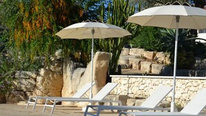 Un área de piscina con sillas de lounge blancas y sombrillas, rodeada de rocas y cactus.