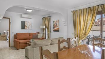 Una sala de estar con un sofá, una mesa de comedor y sillas. Hay dos cuadros en la pared, una ventana con cortinas y un equipo de aire acondicionado.