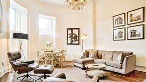 Un salón espacioso con sofá, sillones, una mesa de centro y una araña. En la pared hay cuadros enmarcados y un comedor con mesa y sillas.