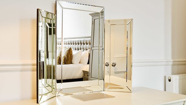 Un dormitorio con un armario espejado, una cama con cabecera capitonada y un aparador blanco.