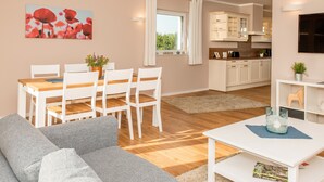 A modern living room with a grey sofa, a dining table with white chairs, a kitchen area with white cabinets, and a painting of red flowers on the wall.