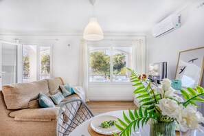 Ein helles Zimmer mit einer Couch, einem Esstisch mit Blumenvase und einem großen Fenster mit Vorhängen.