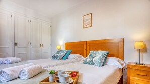 A hotel room with two beds, a wooden headboard, a bedside table with a lamp, and a tray with towels and breakfast items.