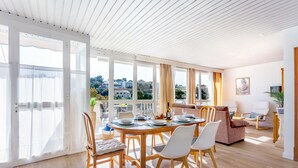 Àmplia zona de dinar amb taula de fusta preparada per a quatre persones, envoltada de cadires blanques. Grans finestres ofereixen vista a la ciutat.
