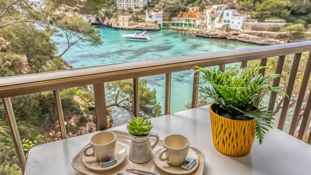 Un balcón con una mesa para dos, una planta en maceta y vistas a un pueblo costero con edificios y un barco.
