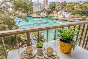 Un balcón con una mesa para dos, una planta en maceta y vistas a un pueblo costero con edificios y un barco.