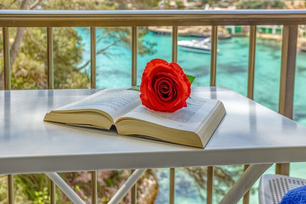 Un libro abierto con una rosa roja sobre una mesa blanca, con vista a un cuerpo de agua turquesa y vegetación.