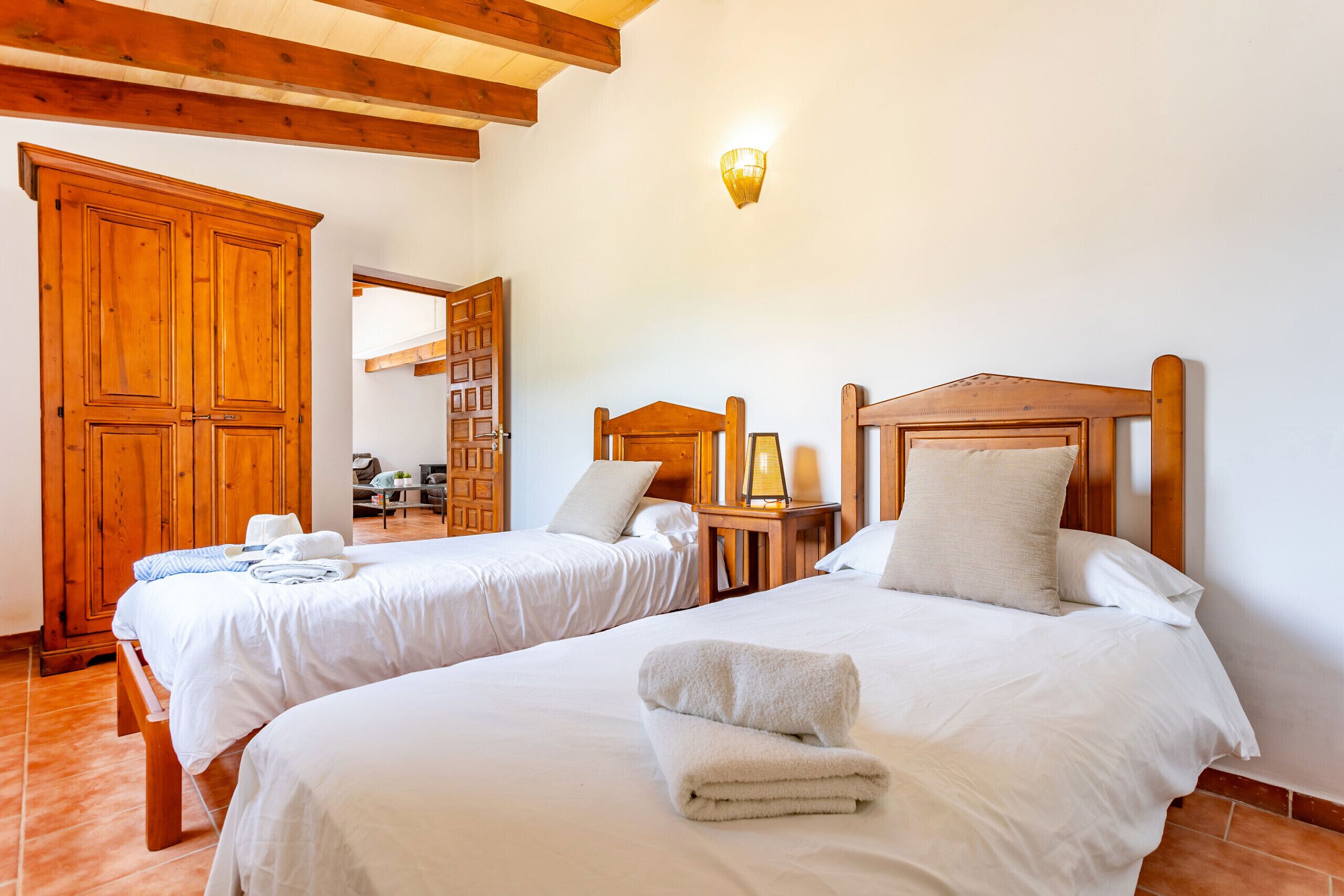 A hotel room with two beds, wooden headboards, a large wardrobe, and a small table with a lamp.