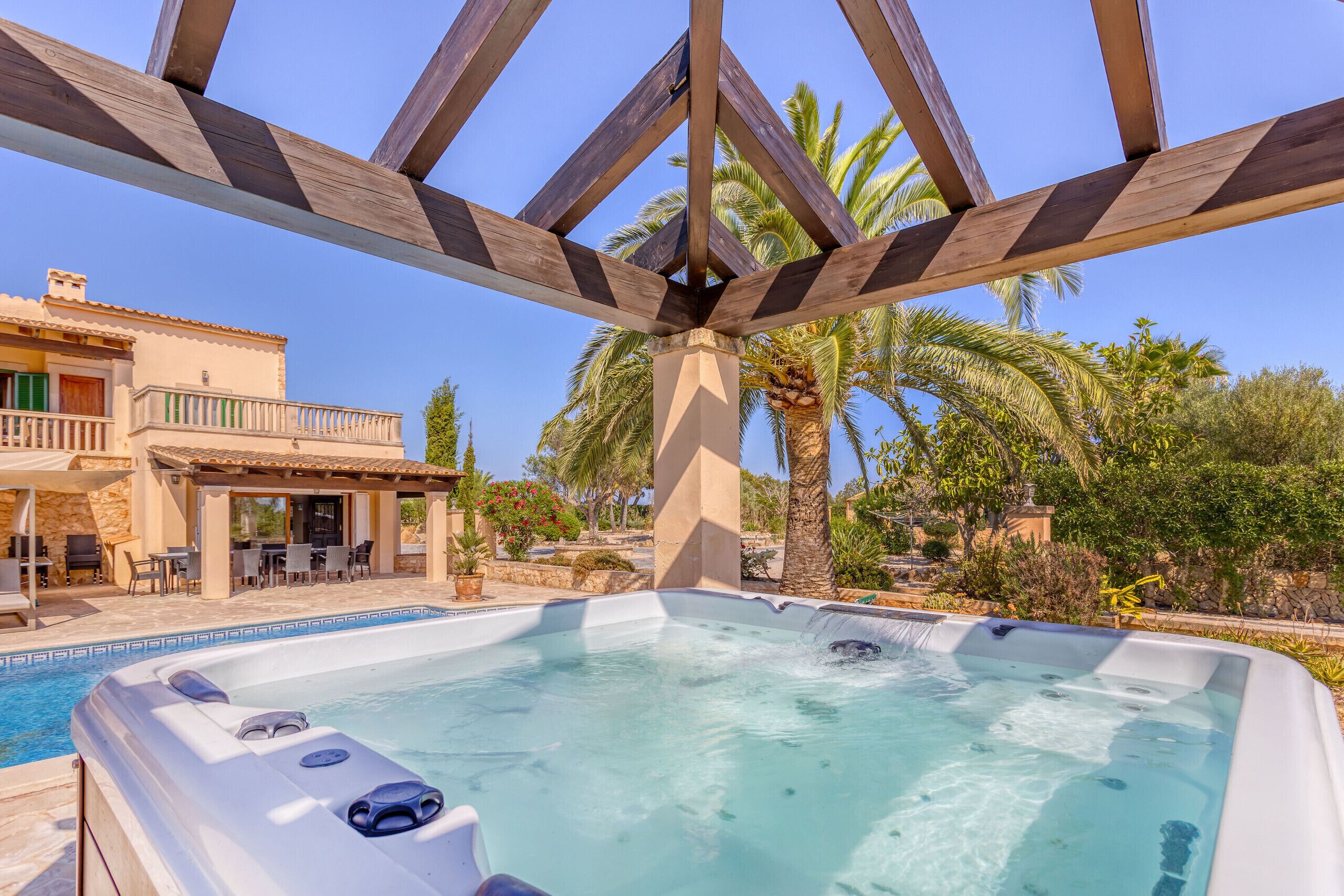 Ein Whirlpool mit einem klaren, blauen Pool, umgeben von einer Holzpergola, Palmen und einem Gebäude mit Balkon.