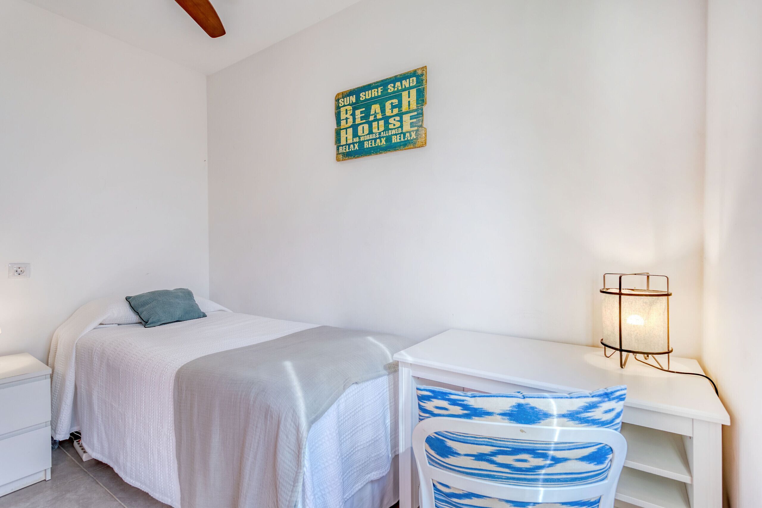 A bedroom with a bed, a desk, and a chair. There is a wall sign that says "SUN SURF SAND BEACH HOUSE RELAX RELAX."