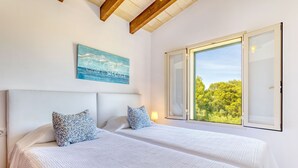 A bedroom with two beds, a large window with a view of greenery, a painting on the wall, and a wooden ceiling beam. - Can Sivinar by dracmallorca (Santanyi)