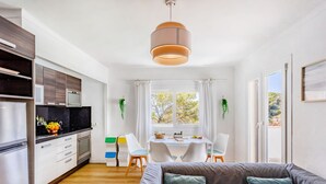 A modern kitchen with white cabinets, a black countertop, and a hanging light fixture. There is a dining area with a table and chairs, and a sofa with cushions.