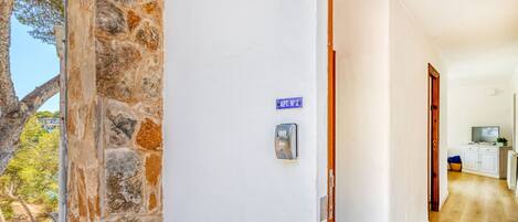 Un pasillo con pared de piedra, una puerta con letrero azul, un dispositivo de seguridad y una zona de cocina con armarios y televisor.