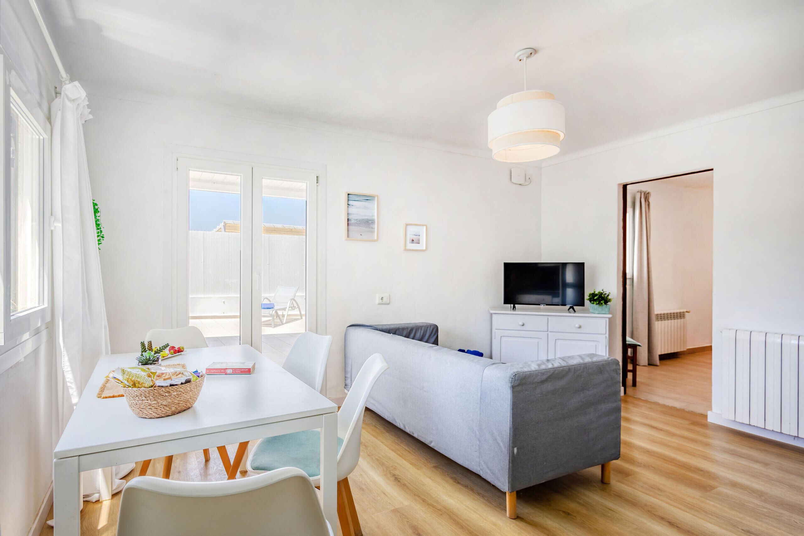 A bright, modern room with a white table, a grey sofa, a TV, and a window with curtains.