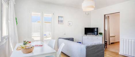 A bright, modern room with a white table, a grey sofa, a TV, and a window with curtains.